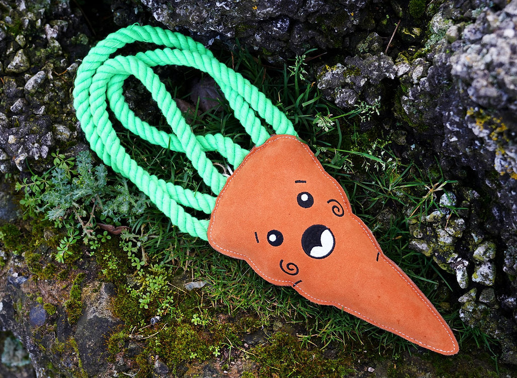 Close-up shot of the orange suede Carrot XL toy with green rope leaves resting on mossy rocks and grass, supplied by Paw Branch.