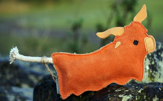 Full view of the orange suede Hamish the Highland Cow toy with rope tail resting on a mossy stone wall, supplied by Paw Branch.