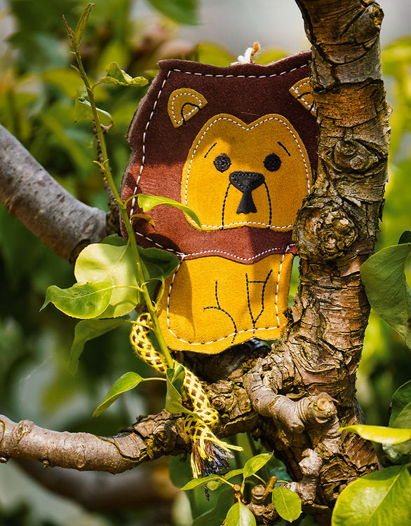 Close-up of the suede Howard the Lion dog toy by Aurora Paws hanging from a green-leaved branch, highlighting the yellow rope detail, supplied by Paw Branch.
