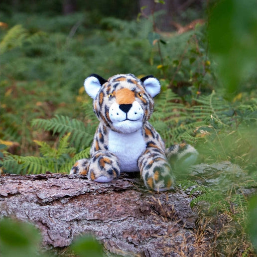 A plush tiger toy sitting on a log in a forest setting with green ferns in the background.