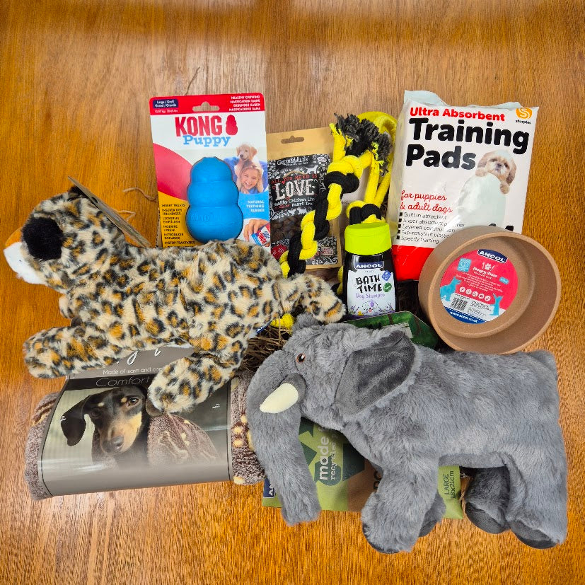 A collection of dog toys and accessories arranged on a wooden floor, including a leopard plush toy, a gray elephant plush toy, a Kong Puppy toy, a training pad package, a dog bowl, and other pet items.