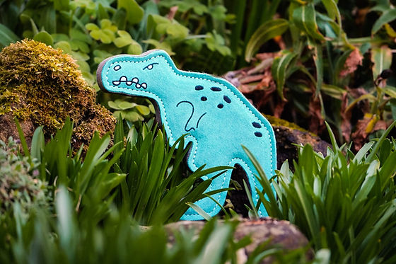 The blue Terry T-Rex dog toy by Aurora Paws standing upright amongst green foliage and mossy rocks in a garden setting, supplied by Paw Branch.